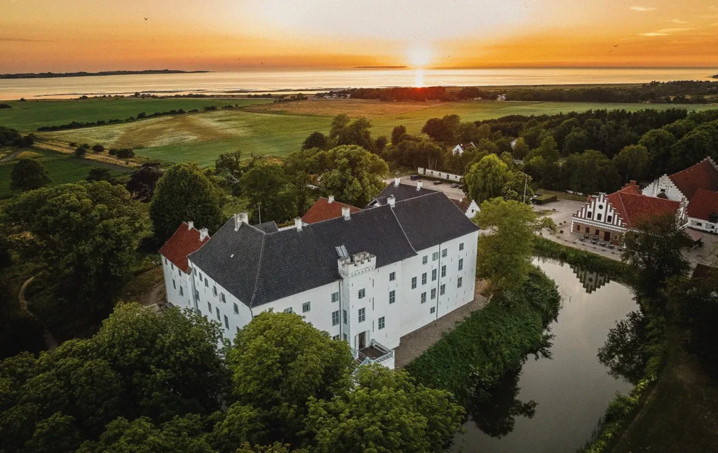 Et billede af Dragholm slot, med solnedgang i horisonten over skv og så. Man kan se de tilhørende stalde og boliger der ligger ved foden af slottet